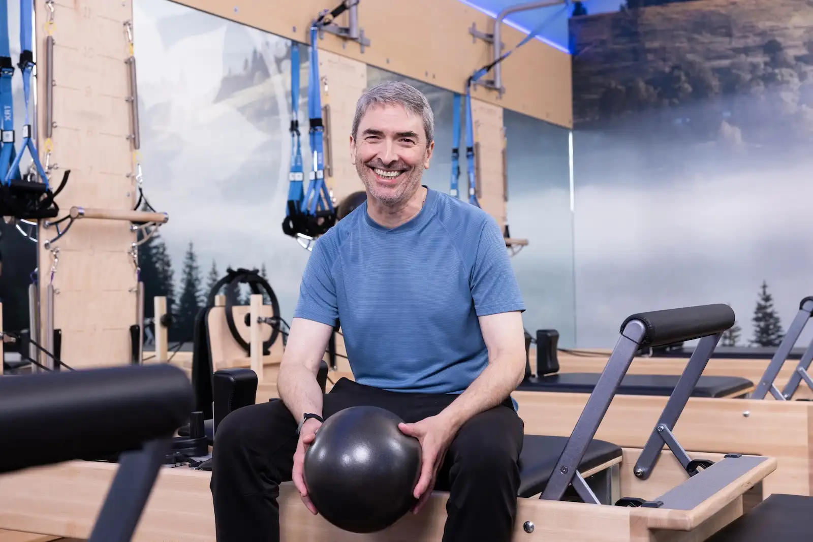 Participante de Club Pilates Bilbao disfrutando del bienestar tras su sesión de Pilates Reformer Señor sentado sobre reformer con pelota en la mano sonriendo después de su rutina en Club Pilates Bilbao Indautxu